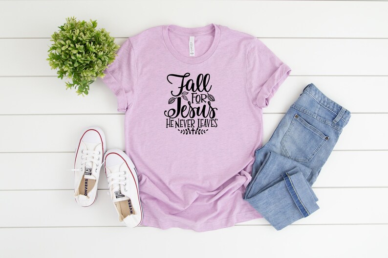 K&ouml;nnte beinhalten: Ein helles lila T-Shirt mit dem Text "Fall for Jesus He Never Leaves" in schwarzer Schrift. Das Shirt liegt flach auf einer wei&szlig;en Holzoberfl&auml;che mit einem Paar wei&szlig;en Turnschuhen und einer blauen Jeans.
