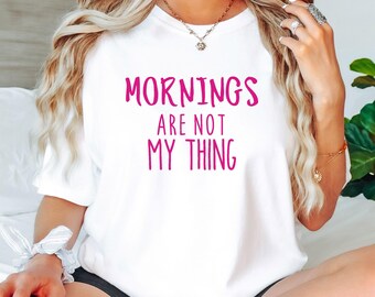 Op de afbeelding: Wit T-shirt met de tekst "Mornings Are Not My Thing" in felroze letters. Het shirt heeft een ronde hals en lijkt gemaakt van een zacht materiaal. Het woord "WHITE" staat in een witte doos onderaan de afbeelding.