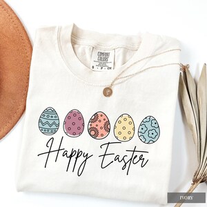 May include: Ivory-colored t-shirt with a "Happy Easter" graphic. The design features five colorful Easter eggs with different patterns. A gold necklace is draped across the shirt. The shirt is labeled "Comfort Colors".
