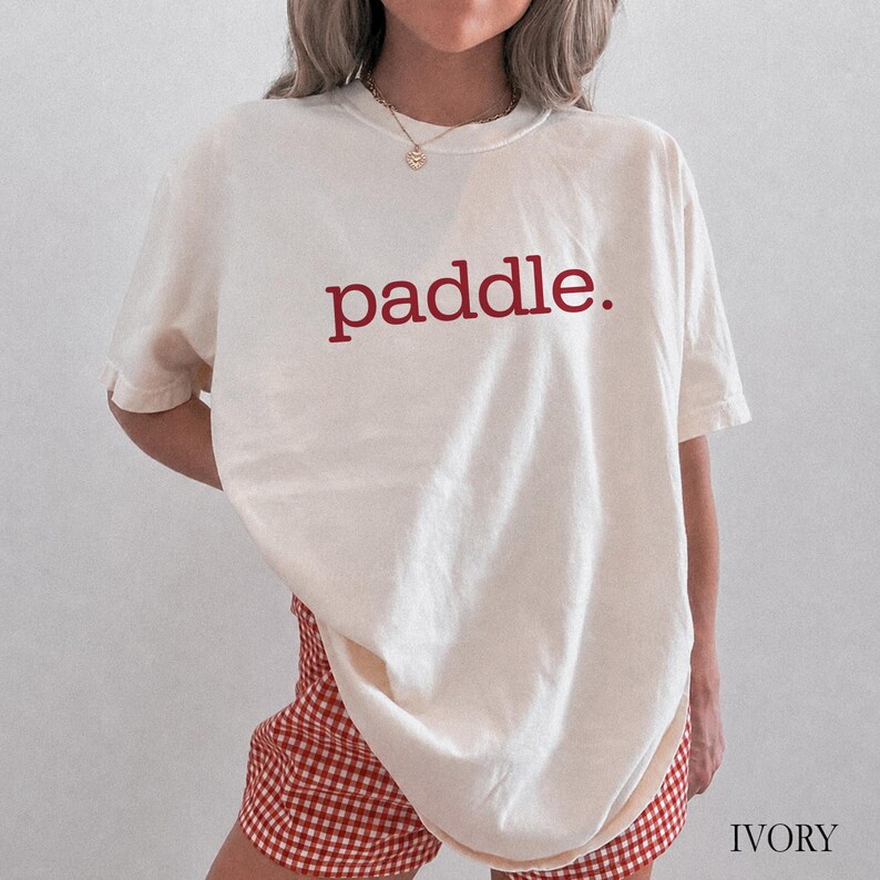 A person wearing an ivory t-shirt with the word paddle printed in red on the chest.