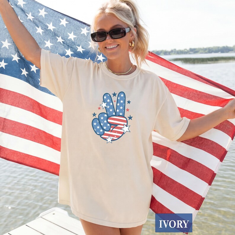 May include: Ivory-colored t-shirt featuring a peace sign design in red, white, and blue, with stars and stripes. The shirt is a relaxed fit. The word "IVORY" is printed at the bottom of the image.