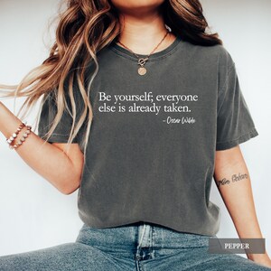 A person wears a dark charcoal grey t-shirt printed with the quote "Be yourself; everyone else is already taken. - Oscar Wilde" in white lettering.