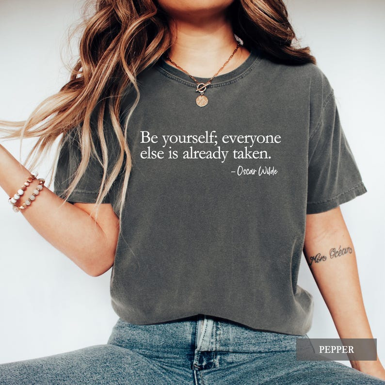 A person wears a dark charcoal grey t-shirt printed with the quote "Be yourself; everyone else is already taken. - Oscar Wilde" in white lettering.