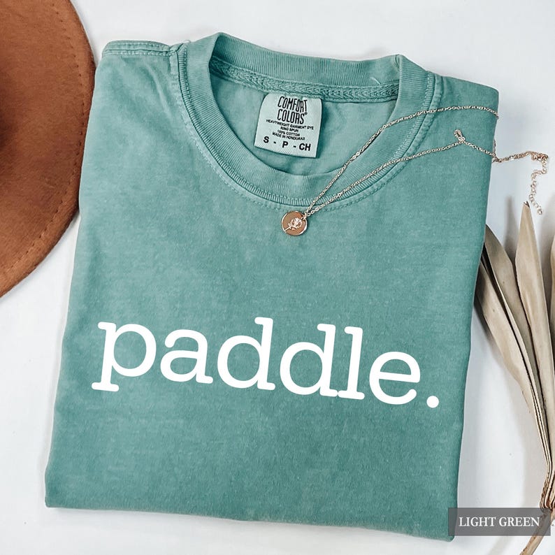 A folded light green t-shirt with the word paddle printed in white across the chest and a necklace resting on the collar.