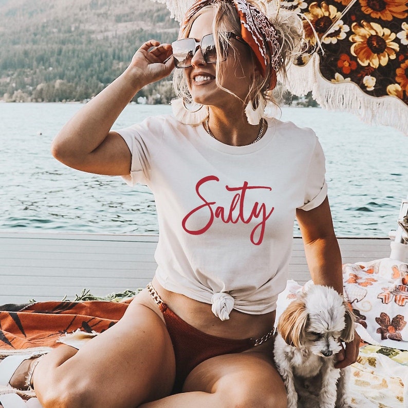 May include: A white t-shirt with the word "Salty" written in red cursive lettering. The shirt is tied at the waist and the model is wearing a brown bikini bottom.