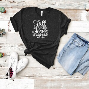 K&ouml;nnte beinhalten: Ein dunkelgraues T-Shirt mit dem Text "Fall for Jesus He Never Leaves" in wei&szlig;en Buchstaben. Das Shirt liegt flach auf einer hellen Holzoberfl&auml;che mit einem Paar wei&szlig;en Sneakers und einer hellblauen Jeans.