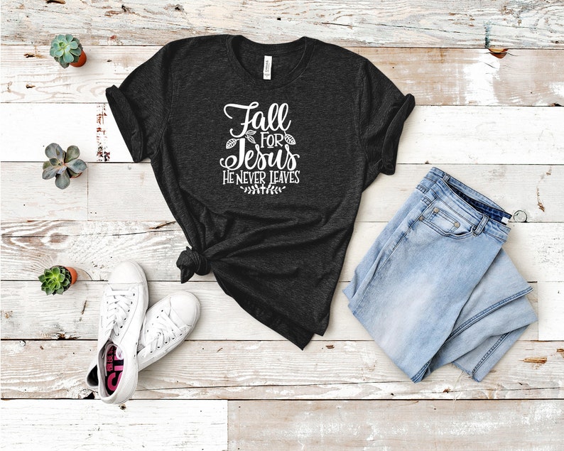 K&ouml;nnte beinhalten: Ein dunkelgraues T-Shirt mit dem Text "Fall for Jesus He Never Leaves" in wei&szlig;en Buchstaben. Das Shirt liegt flach auf einer hellen Holzoberfl&auml;che mit einem Paar wei&szlig;en Sneakers und einer hellblauen Jeans.