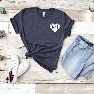 May include: A dark blue t-shirt with a white heart design featuring a baseball inside. The shirt is laid out on a white wooden surface with a pair of white sneakers and a pair of light blue jeans.