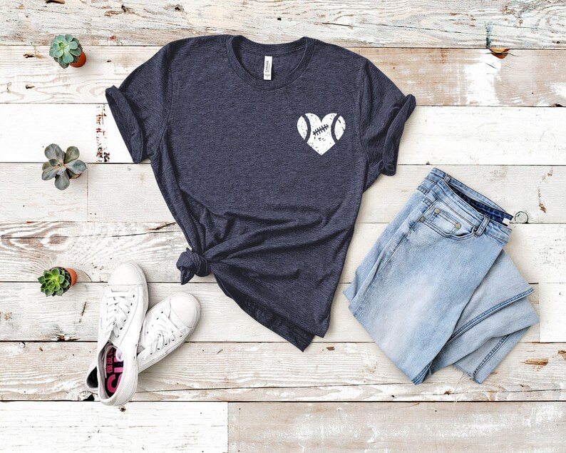 May include: A dark blue t-shirt with a white heart design featuring a baseball inside. The shirt is laid out on a white wooden surface with a pair of white sneakers and a pair of light blue jeans.