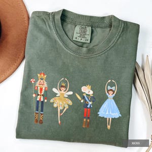 May include: A moss green t-shirt featuring a Nutcracker-themed design. The graphic includes a nutcracker, a ballerina, a mouse king, and another ballerina. The shirt has a crew neck and short sleeves. The tag reads "Comfort Colors".