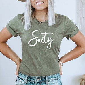 May include: A woman wearing a green t-shirt with the word "Salty" written in white cursive lettering.