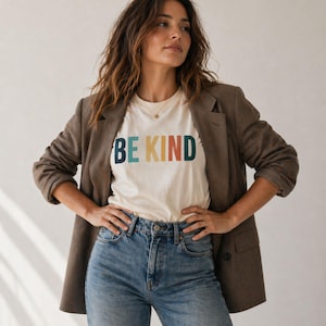 May include: A cream-colored t-shirt with the words "BE KIND" in colorful block letters. The person is wearing a brown blazer and blue jeans. The overall style is casual and comfortable.