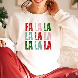 May include: White sweatshirt with a festive Christmas design featuring the text "Fa La La La La La La La" in red, pink, green, and blue with a distressed look.