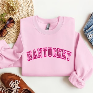 Puede incluir: Una sudadera de cuello redondo en color rosa claro con la palabra NANTUCKET impresa en una fuente de estilo universitario de color rosa oscuro en el pecho. La prenda tiene mangas largas, cuello acanalado y una etiqueta de marca visible.