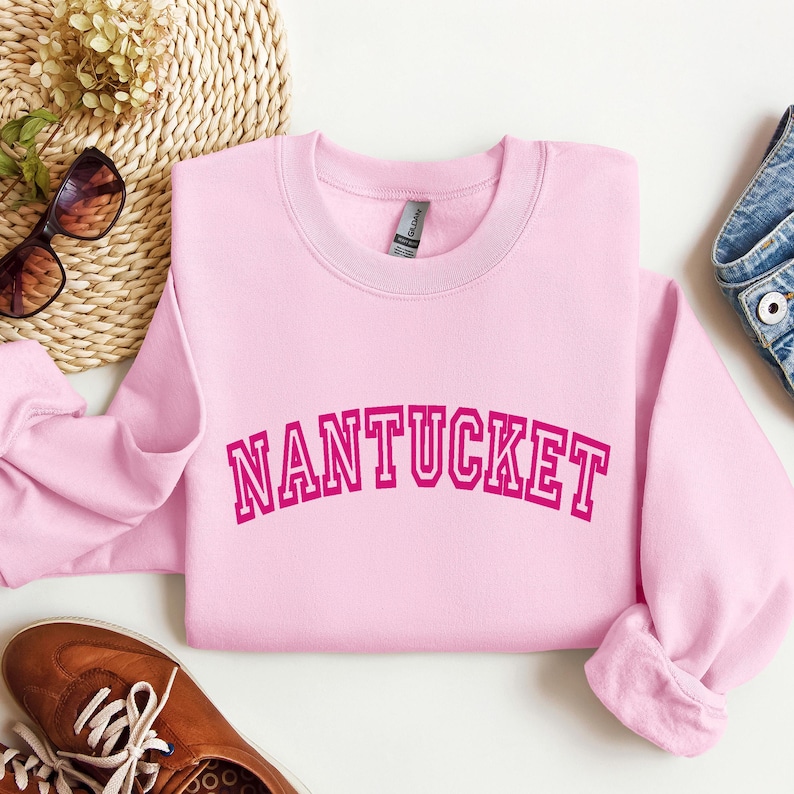 Puede incluir: Una sudadera de cuello redondo en color rosa claro con la palabra NANTUCKET impresa en una fuente de estilo universitario de color rosa oscuro en el pecho. La prenda tiene mangas largas, cuello acanalado y una etiqueta de marca visible.