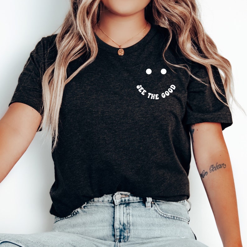 K&ouml;nnte beinhalten: Ein dunkelgraues T-Shirt mit einem wei&szlig;en Aufdruck, der "See the good" sagt, mit zwei wei&szlig;en Punkten &uuml;ber dem Text, die ein Smiley-Gesicht bilden.