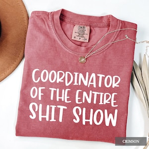 以下が含まれることがあります： 「COORDINATOR OF THE ENTIRE SHIT SHOW」の白文字がプリントされたクリムゾンカラーのTシャツ。クルーネックで、右上に小さなタグが付いています。ペンダント付きのシルバーネックレスがTシャツの上にかけられています。
