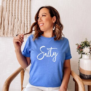 May include: A woman wearing a light blue t-shirt with the word "Salty" written in white cursive lettering.