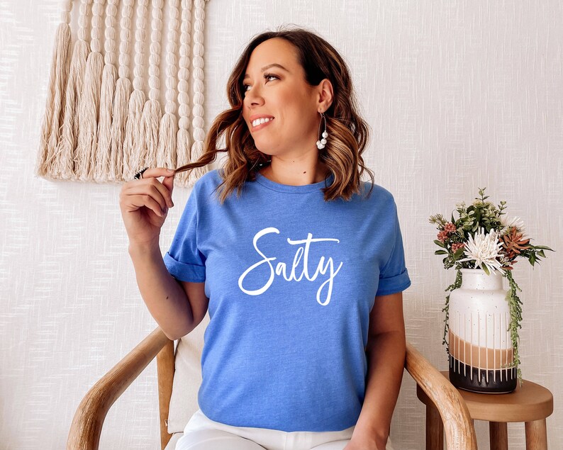 May include: A woman wearing a light blue t-shirt with the word "Salty" written in white cursive lettering.