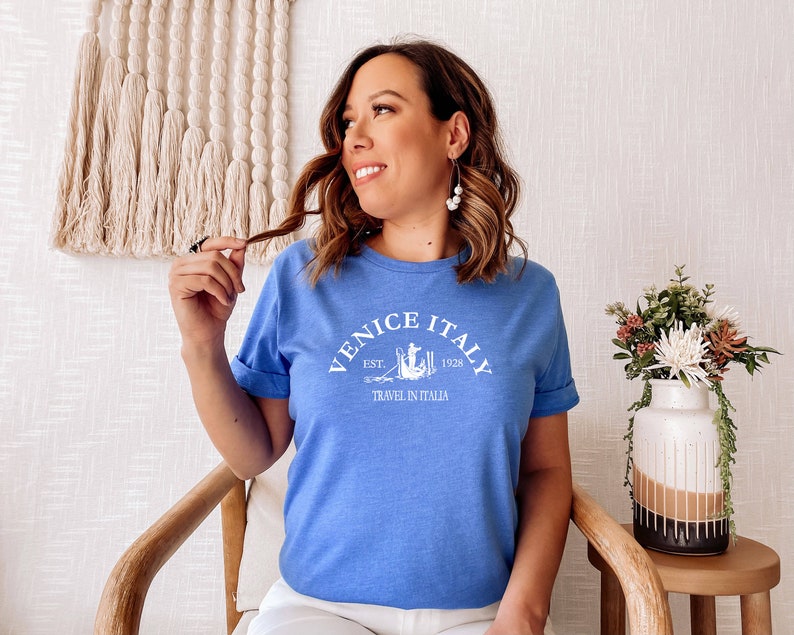 May include: A blue t-shirt with white text that reads "Venice Italy Est. 1928 Travel in Italia". The text is in a vintage style font and includes a small graphic of a gondola.