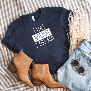May include: A navy blue t-shirt with white text that reads "I was normal 2 kids ago". The shirt is laid flat on a striped surface with brown boots, blue jeans, and dried flowers.