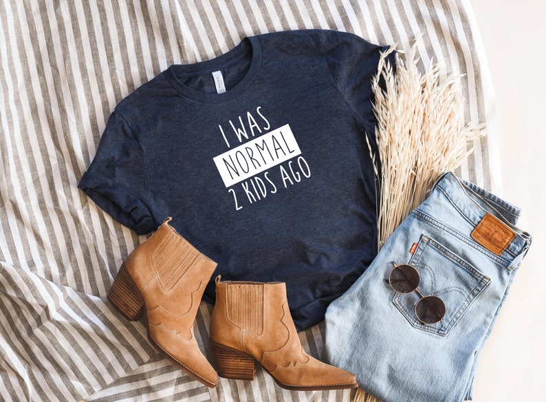 May include: A navy blue t-shirt with white text that reads "I was normal 2 kids ago". The shirt is laid flat on a striped surface with brown boots, blue jeans, and dried flowers.