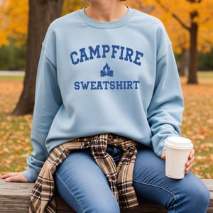 A person wearing a light blue crewneck sweatshirt with "CAMPFIRE SWEATSHIRT" in dark blue text and a small campfire icon, sits outdoors.
