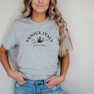 May include: A gray t-shirt with a black graphic design that says "Venice Italy Est. 1928 Travel in Italia".