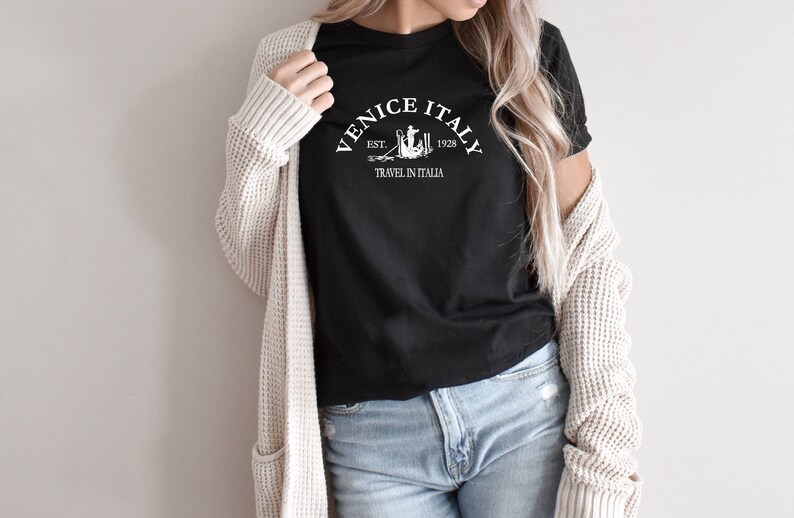 May include: A black t-shirt with a white graphic design that says "Venice Italy Est. 1928 Travel in Italia".