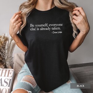 A person wears a black t-shirt printed with the quote "Be yourself; everyone else is already taken. - Oscar Wilde" in white lettering.