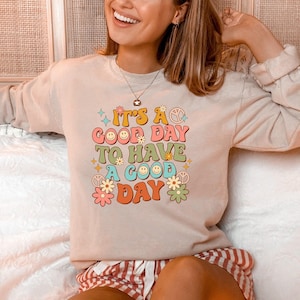 Könnte beinhalten: Ein hellbeiger Pullover mit dem Text "It's a good day to have a good day" in einer Retro-Schriftart mit bunten Blumen und Peace-Zeichen.