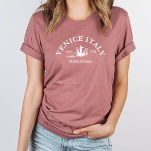 May include: A mauve t-shirt with a white graphic design that says "Venice Italy Est. 1928 Travel in Italia".