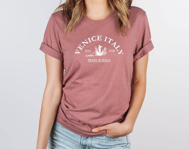 May include: A mauve t-shirt with a white graphic design that says "Venice Italy Est. 1928 Travel in Italia".