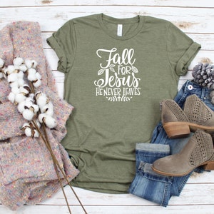 K&ouml;nnte beinhalten: Ein gr&uuml;nes T-Shirt mit dem Text "Fall for Jesus He Never Leaves" in wei&szlig;en Buchstaben. Das Shirt ist mit einer blauen Jeans, braunen Stiefeln und einem rosa-wei&szlig;en Pullover gestylt.
