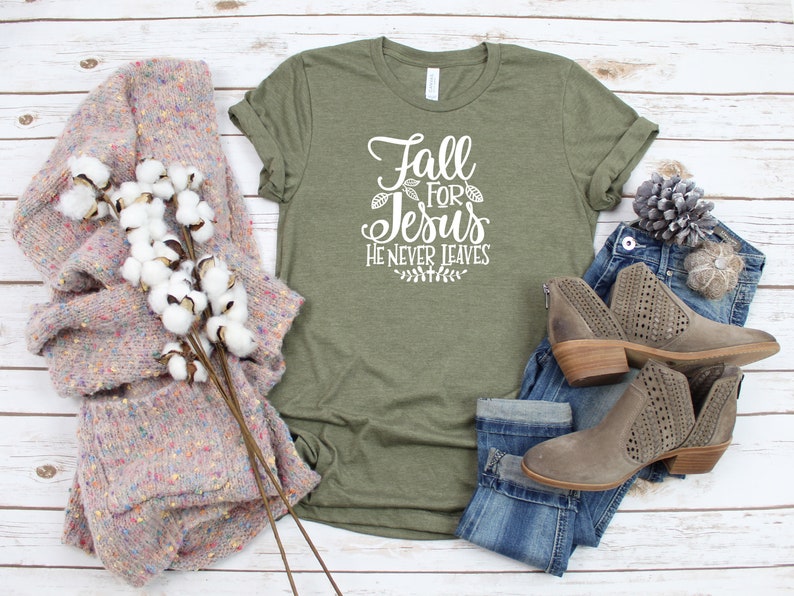 K&ouml;nnte beinhalten: Ein gr&uuml;nes T-Shirt mit dem Text "Fall for Jesus He Never Leaves" in wei&szlig;en Buchstaben. Das Shirt ist mit einer blauen Jeans, braunen Stiefeln und einem rosa-wei&szlig;en Pullover gestylt.