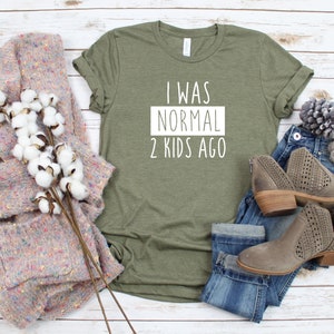 May include: A green t-shirt with the text "I was normal 2 kids ago" in white lettering. The shirt is laid flat on a white wooden surface with a pair of jeans, a sweater, and boots.