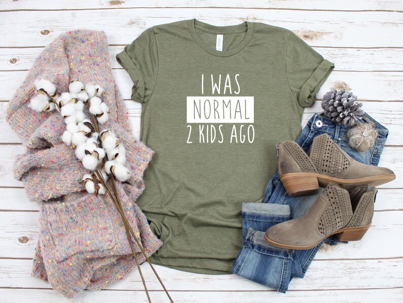 May include: A green t-shirt with the text "I was normal 2 kids ago" in white lettering. The shirt is laid flat on a white wooden surface with a pair of jeans, a sweater, and boots.
