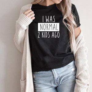 May include: A black t-shirt with white text that reads "I was normal 2 kids ago". The shirt is being worn by a person with long blonde hair and a white cardigan.