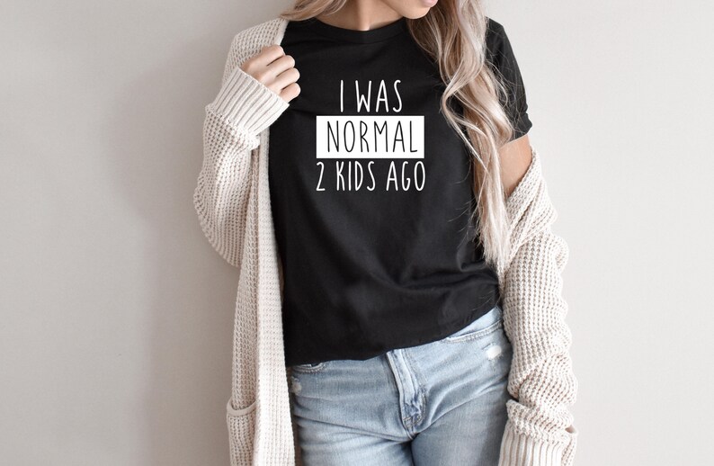 May include: A black t-shirt with white text that reads "I was normal 2 kids ago". The shirt is being worn by a person with long blonde hair and a white cardigan.