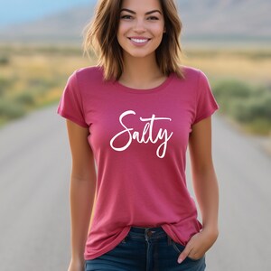 May include: A woman wearing a pink t-shirt with the word "Salty" written in white cursive script.