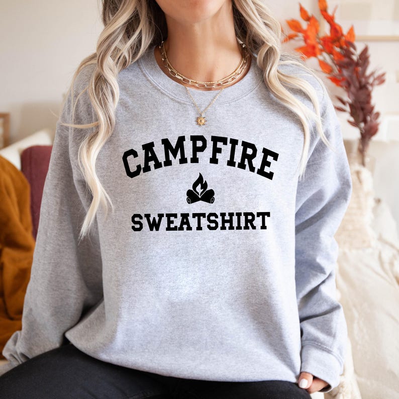A person wearing a light gray crewneck sweatshirt with "CAMPFIRE SWEATSHIRT" in black text and a small campfire icon, stands indoors.