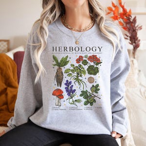 May include: Gray sweatshirt with a graphic print of various plants and the text "HERBOLOGY HOGWARTS SCHOOL OF WITCHCRAFT AND WIZARDRY" with the names of each plant listed below.