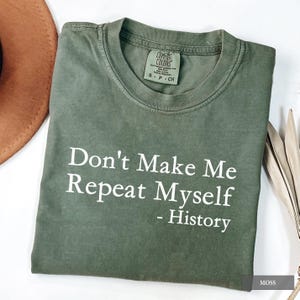 Op de afbeelding: Een mosgroen t-shirt met de witte tekst "Don't Make Me Repeat Myself - History". Het shirt is opgevouwen en heeft een Comfort Colors-label. Een bruine hoed en gedroogde bladeren zijn gedeeltelijk zichtbaar.
