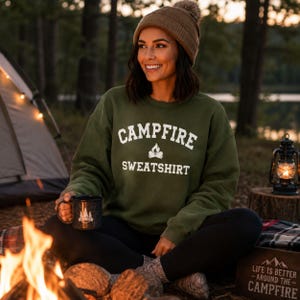 A woman sits by a campfire, wearing an olive green crewneck sweatshirt with "CAMPFIRE SWEATSHIRT" in white text and a small campfire icon.