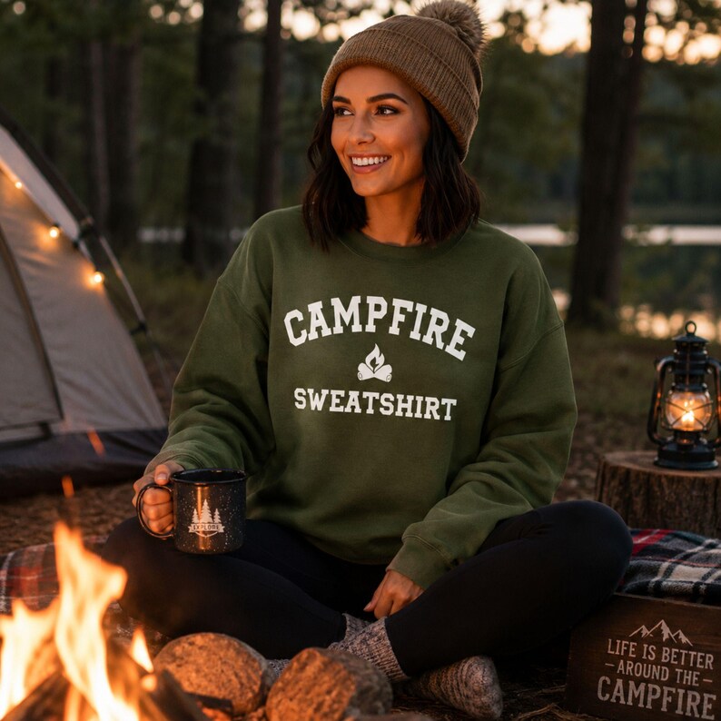 A woman sits by a campfire, wearing an olive green crewneck sweatshirt with "CAMPFIRE SWEATSHIRT" in white text and a small campfire icon.