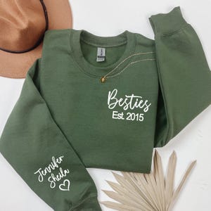 以下が含まれることがあります： 前面に「Besties Est 2015」の文字、袖に「Jennifer Sheila」とハートの文字が入った緑色のスウェットシャツです。