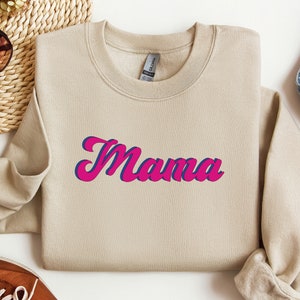 May include: A tan sweatshirt with the word "Mama" printed in pink and blue letters in a retro style font.