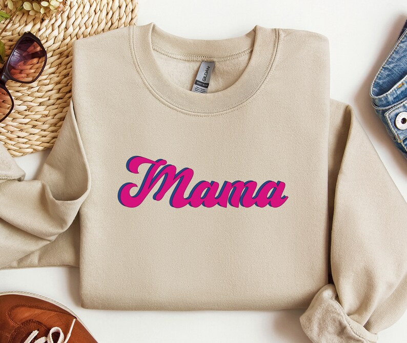 May include: A tan sweatshirt with the word "Mama" printed in pink and blue letters in a retro style font.