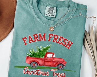 May include: Light green t-shirt with the words "FARM FRESH" in red above a red pickup truck carrying a green Christmas tree. The words "Christmas Trees" are below the truck in red. A silver necklace with a pendant is on the shirt.