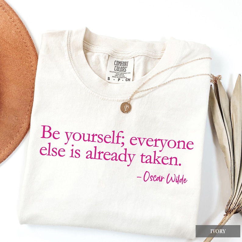 An ivory Comfort Colors t-shirt printed with the quote "Be yourself; everyone else is already taken. - Oscar Wilde" in dark pink lettering.